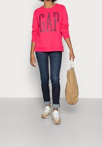 Felpa rosa con logo "GAP", abbinata a jeans skinny scuri e sneakers beige. Porta una borsa color cioccolato, con una vestibilità rilassata e collo a giro.