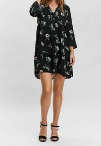 Svart blommig klänning med V-ringning, långa ärmar och ledad design med vita och gröna blomster mönster. Bärs med svarta klackskor med rem runt ankeln.