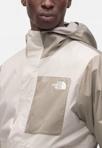 Gros plan d'une personne portant une veste à capuche beige clair et taupe avec le logo The North Face sur la poche gauche de la poitrine.