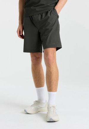 Mand iført sorte shorts, hvide sokker og hvide sportssko, stående med den ene hånd i lommen mod en ensfarvet baggrund.