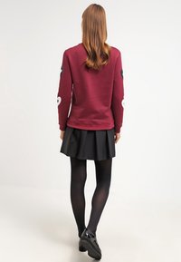 Burgundowa bluza z długim rękawem z czarno-białymi akcentami w kształcie serc, w połączeniu z czarną spódnicą plisowaną i czarnymi rajstopami.