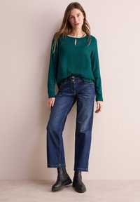Blusa a maniche lunghe color turchese con dettaglio a chiave, abbinata a jeans a gamba larga blu scuro e stivaletti neri su uno sfondo neutro.