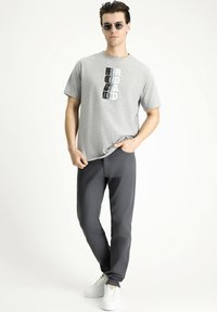 Camiseta gris de manga corta con un estampado gráfico, combinada con pantalones gris oscuro y zapatillas blancas. Las gafas de sol completan el conjunto.