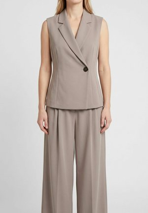 Vestă taupe fără mâneci, croită, cu închidere cu un singur nasture, purtată împreună cu pantaloni largi asortați pe o persoană în picioare.