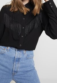 Chemise noire boutonnée avec un design à franges sur la poitrine, associée à un jean taille haute bleu clair. Texture de tissu lisse.