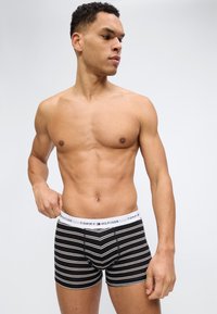 Άνδρας χωρίς πουκάμισο που διορθώνει μαύρα και άσπρα ριγέ μποξεράκια Tommy Hilfiger, κοιτάζοντας προς τα αριστερά του πάνω σε απλό γκρι φόντο.