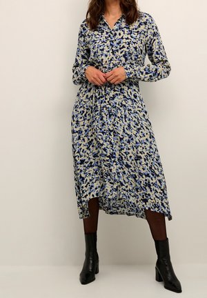 Femme portant une robe midi à manches longues boutonnée avec un imprimé abstrait bleu, noir et beige, associée à des bottines noires à talons.