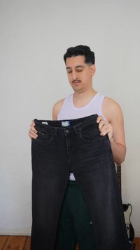 Άνδρας με λευκή αμάνικη μπλούζα κρατάει ένα ζευγάρι μεγάλα μαύρα τζιν παντελόνια, σταθείς μέσα σε κλειστό χώρο με μια κιθάρα να φαίνεται μερικώς πίσω του.