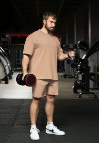 T-shirt et short de sport beiges, tenant des haltères noirs avec des accents rouges, dans un cadre de salle de sport avec des équipements d'exercice en arrière-plan.