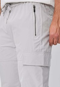 Pantalon cargo gris clair avec poche zippée sur la hanche et poche à rabat sur la cuisse, porté par une personne dont la main droite est visible.