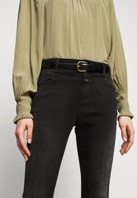Jeans negros de corte slim, combinados con una blusa verde oliva clara con puños con plisado y un cinturón negro con un detalle de hebilla dorada.