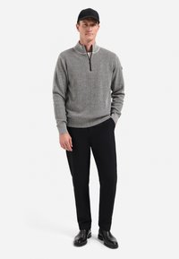 Man in grijze rits trui, zwarte broek, zwarte pet en zwarte laarzen die met één hand in zijn zak staat tegen een effen witte achtergrond.