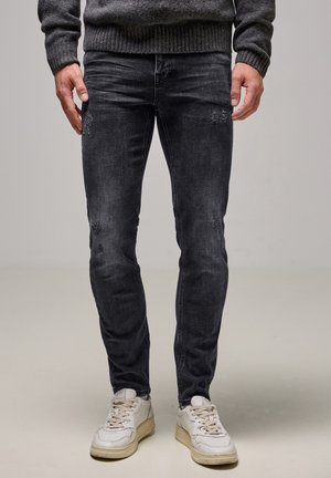 Uomo che indossa jeans skinny neri consumati, un maglione grigio scuro e sneakers bianche, in piedi su uno sfondo chiaro e semplice.