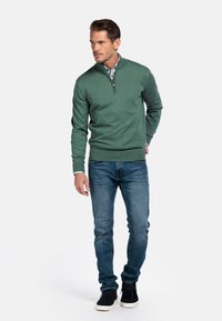 Groene pullover trui met een halve rits kraag, gecombineerd met blauwe denim jeans en zwarte sneakers. Heeft een gestructureerde stof en ribbels aan de mouwen.