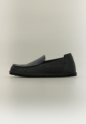 Mocassin noir sans lacets avec dessus cousu et semelle en caoutchouc plate vu de côté sur fond neutre.