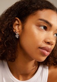 Pendientes de plata con diversas formas geométricas, incluyendo un corazón, cristales transparentes y acentos metálicos, usados por un modelo con cabello rizado.