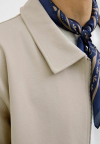 Beige zip-up jacket met een platte kraag, textuurstof; gedragen met een marineblauwe sjaal met gouden bladeren patronen.