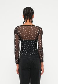 Schwarzes Mesh-Top mit Polka-Dots und langen Ärmeln, das eine taillierte Passform mit Raffung an der Taille und einem gewellten Ausschnitt aufweist.