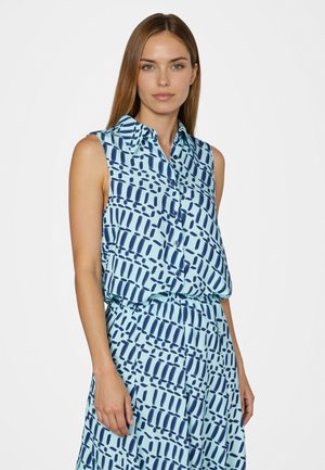Frau trägt ärmelloses, mit Kragen und Knöpfen versehenes blaues Oberteil und passenden Rock mit abstraktem marineblauem geometrischem Muster auf hellblauem Hintergrund.