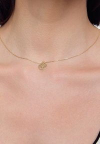 Złoty naszyjnik z okrągłym wisiorkiem z motywem drzewa, zawieszony na delikatnym łańcuszku; gładka, wypolerowana tekstura.