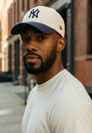 Bärtiger Mann, der eine weiße und marineblaue Mütze sowie ein weißes Hemd trägt und auf einer Stadtstraße mit Backsteingebäuden zur Kamera blickt.