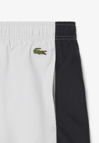 Zwart-witte zwemshorts met een elastische tailleband en een groene krokodillenlogo ingeborduurd aan de linkerkant. Glad, lichtgewicht materiaal.