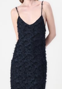 Robe bleu marine avec un décolleté en V, des fines bretelles et un tissu texturé présentant un motif d'accents surélevés et effilochés.