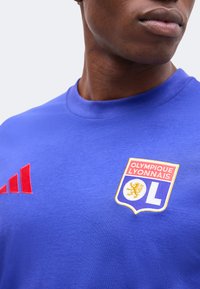 Άνδρας που φοράει μπλε μπλούζα Olympique Lyonnais με κόκκινο λογότυπο Adidas και έμβλημα ομάδας με λιοντάρι στο στήθος.