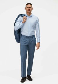 Lichtblauw overhemd, op maat gemaakte lichtblauwe broek, donker colbert over de schouder gedrapeerd, zwarte schoenen, staande houding, tegen een witte achtergrond.