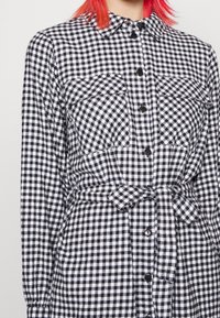 Vestido camisa de cuadros vichy en blanco y negro con mangas largas, dos bolsillos frontales, cierre de botones y detalle de cintura anudada. Fabricado con tela de patrón a cuadros.