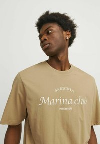 Kamelinės spalvos medvilnės marškinėliai su centriniu baltu užrašu "Sardinia Marina club PREMIUM", trumpomis rankovėmis ir laisvu apdailos stiliumi.
