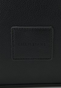 Black textured leather wallet featuring a rectangular patch with debossed brand name "CALL IT SPRING." Minimalist design with black stitching.