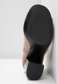 Bottine beige en suède inclinée contre une surface blanche, montrant sa semelle en caoutchouc noir texturé et son talon bloc épais.
