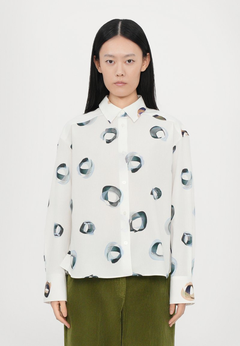 Blusa con botones de fondo blanco, que presenta patrones circulares coloridos en azul, verde y marrón. Mangas largas, corte relajado, textura suave.