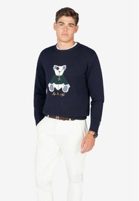 Suéter de punto azul marino con un diseño de oso en la parte frontal, con acentos en blanco, verde y azul. Puños acanalados, cuello redondo y corte relajado.
