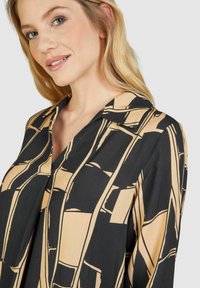 Schwarze und beige gemusterte Bluse mit kragenähnlichem Ausschnitt. Der Stoff hat eine glatte Textur und weist ein geometrisches Design mit klaren Formen auf.