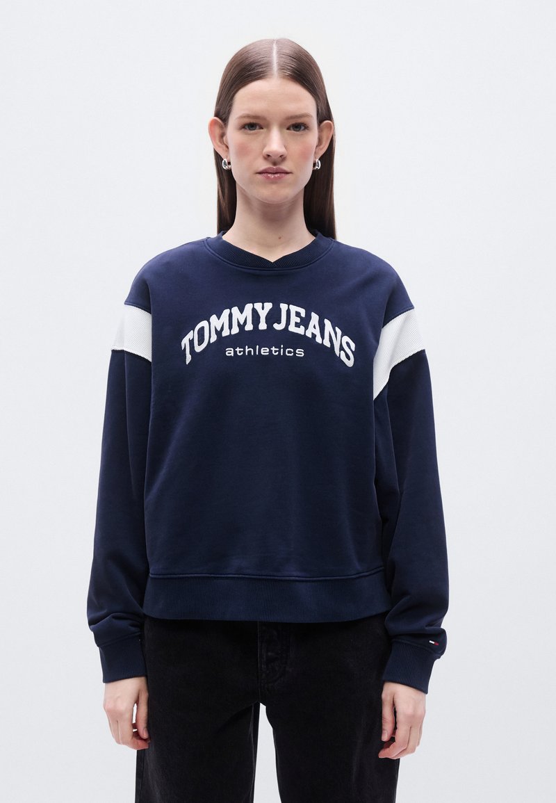 Μπλε σκούρο φούτερ με λευκό κείμενο "TOMMY JEANS athletics", με αντίθετες λευκές λεπτομέρειες στους ώμους και άνετη εφαρμογή. Ριμπ λαιμός και μανσέτες.