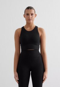 Melnā pieguļošā sporta krūšturī un augstās jostasvietas legingos, gluda auduma ar minimālistisku dizainu, ar apaļu kakla izgriezumu un bez redzamām logotipiem.