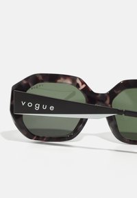 Ochelari de soare octagonali cu ramă din fâșii de țestoasă întunecată, lentile verzi și un braț metalic negru cu branding "vogue" în text alb.