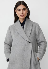 Kvinna med långt mörkt hår iklädd en ljusgrå ullrock med breda slag och händerna i fickorna, mot en enkel vit bakgrund.