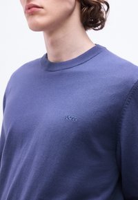 Jeune personne portant un pull bleu à col rond avec "BOSS" brodé sur la poitrine, visible du cou jusqu'au milieu du torse.