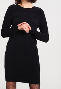 Svart stickad klänning med långa ribbade ärmar, rund halsringning och en figursydd passform. Har en subtil logotyp på bröstet. Strukturerade tygdetaljer.