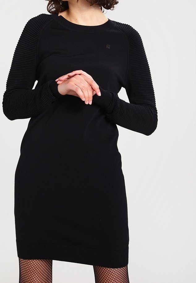 Svart stickad klänning med långa ribbade ärmar, rund halsringning och en figursydd passform. Har en subtil logotyp på bröstet. Strukturerade tygdetaljer.