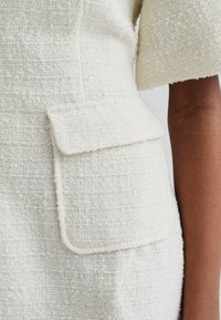 Tissu blanc texturé avec un motif tissé, comportant une poche à rabat sur le côté gauche et un bord légèrement effiloché le long des manches.
