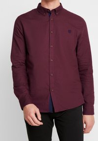 Man in een bordeauxrode, lange mouwen overhemd met een klein logo op de linkerborst en zwarte broek.