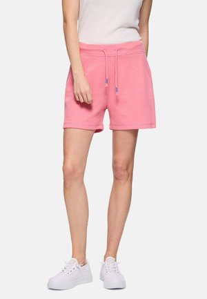 Mujer vestida con pantalones cortos rosa con cordón, camiseta blanca sin mangas y zapatillas blancas, de pie contra un fondo claro y uniforme.