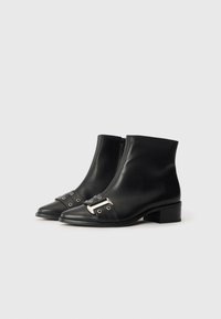 Botas de tornozelo em couro preto com fecho de zip, bico afinado, detalhe de fivela prateada e pequeno salto bloco. Textura suave e design minimalista.