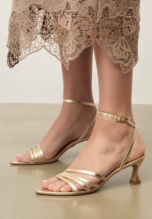Nohy v zlatých sandáloch na nízkom opätku so strapcami a prackami na členkoch, spojené s béžovými čipkovanými šatami s kvetinovými výrezmi.