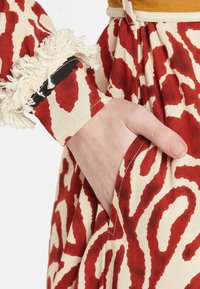 Tessuto a righe rosso e beige con forme astratte, caratterizzato da un accento di frangia testurizzata sul polsino e una mano che riposa in una tasca laterale.