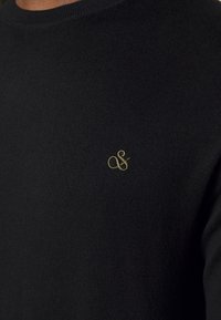 Suéter negro de punto con cuello redondo y un pequeño logotipo bordado en oro en el pecho. La tela parece suave con un ligero brillo.